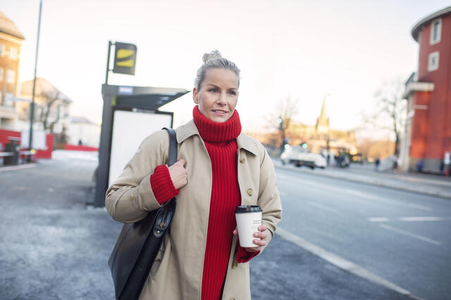 Kvinne i rød høyhalsetgenser og frakk står med en kaffekopp foran holdeplass ved Studentersamfundet.