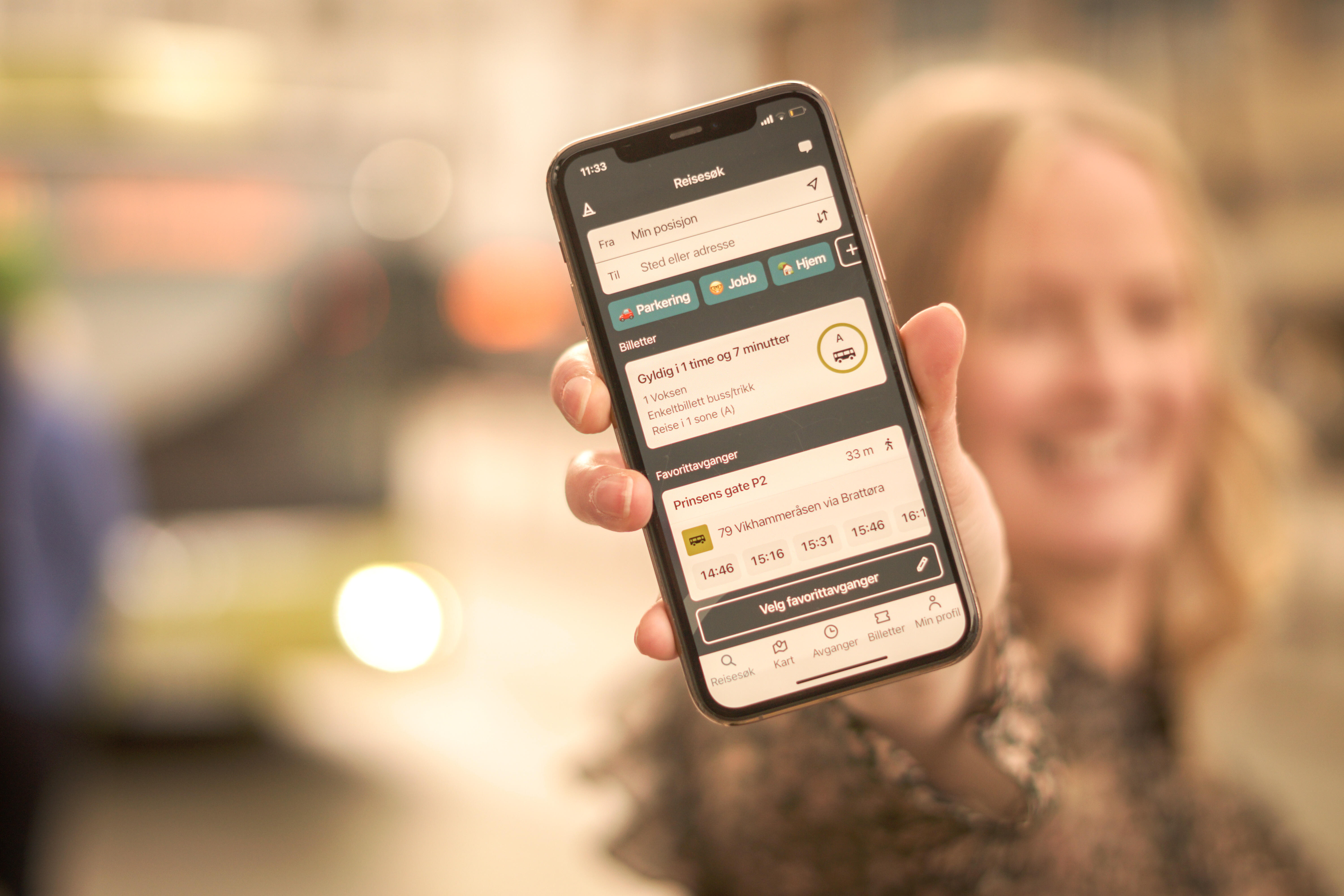Ung kvinne holder smilende opp en telefon med AtB-appen åpen på skjermen. En buss kan skimtes i bakgrunnen.