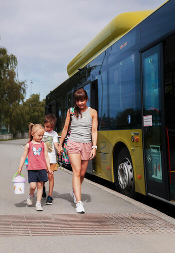 To barn og en forelder i sommerklær på vei inn i buss.
