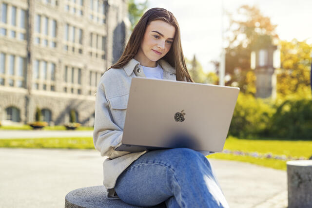 Kvinne med langt hår med PC på fanget og studiested i bakgrunnen.