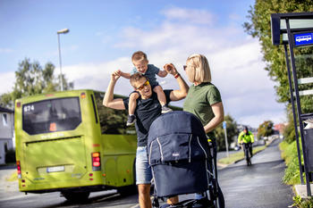 Tre personer med barnevogn foran seg, og grønn buss i bakgrunnen